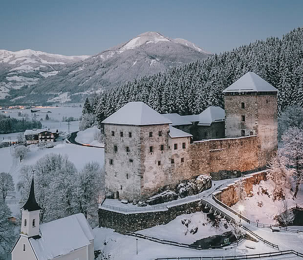 Burg Kaprun | 1,1 km die beleuchtete Burg Kaprun mit der Kapelle in der mit Schnee bedeckten Landschaft bei Dämmerung.