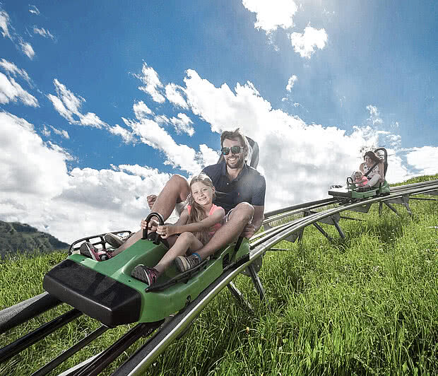 Maisi Flitzer | 260 m Vater und Tochter fahren lachend mit einem grünen Schlitten eine Sommerrodelbahn hinab. Im Hintergrund ist ein weiteres Paar auf der Bahn sichtbar. Die Strecke verläuft durch grüne Wiesen unter einem strahlend blauen Himmel mit weißen Wolken.